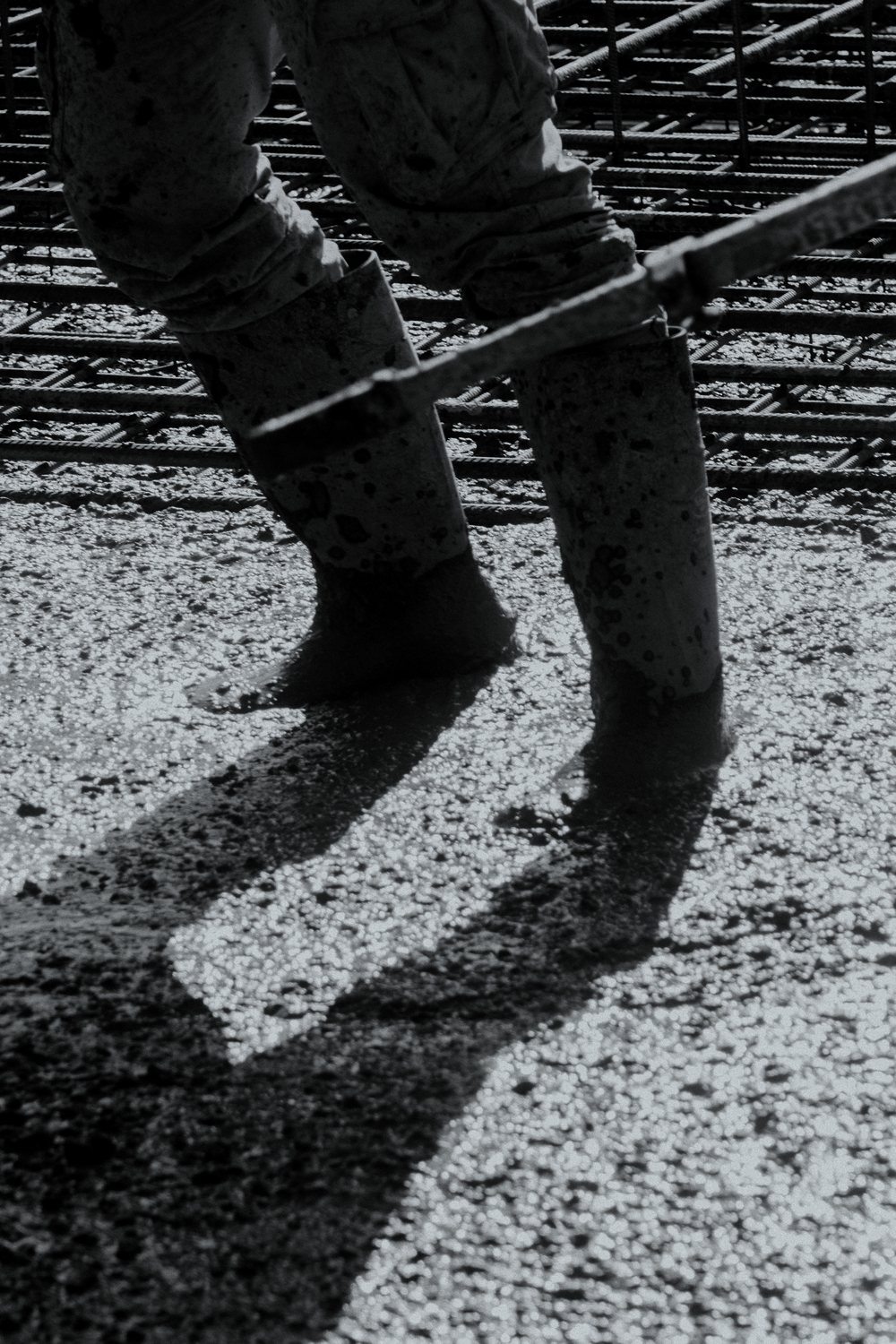 A worker standing in wet concrete over a rebar mat, boots buried past the ankle, pulling a screed rail across the pour.