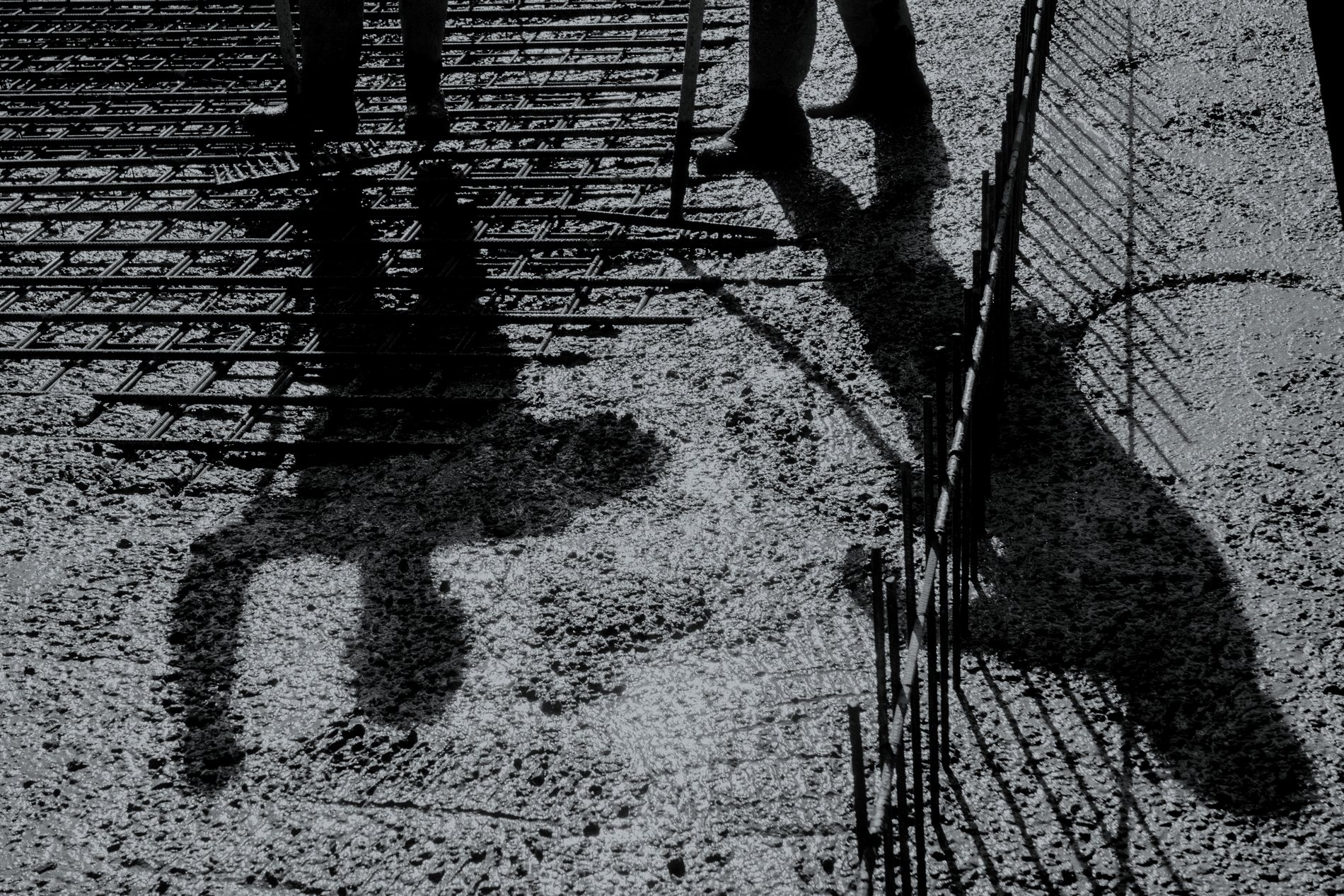 Fresh concrete being placed over a rebar mat, with the shadows of two workers and a rake falling across the wet surface.