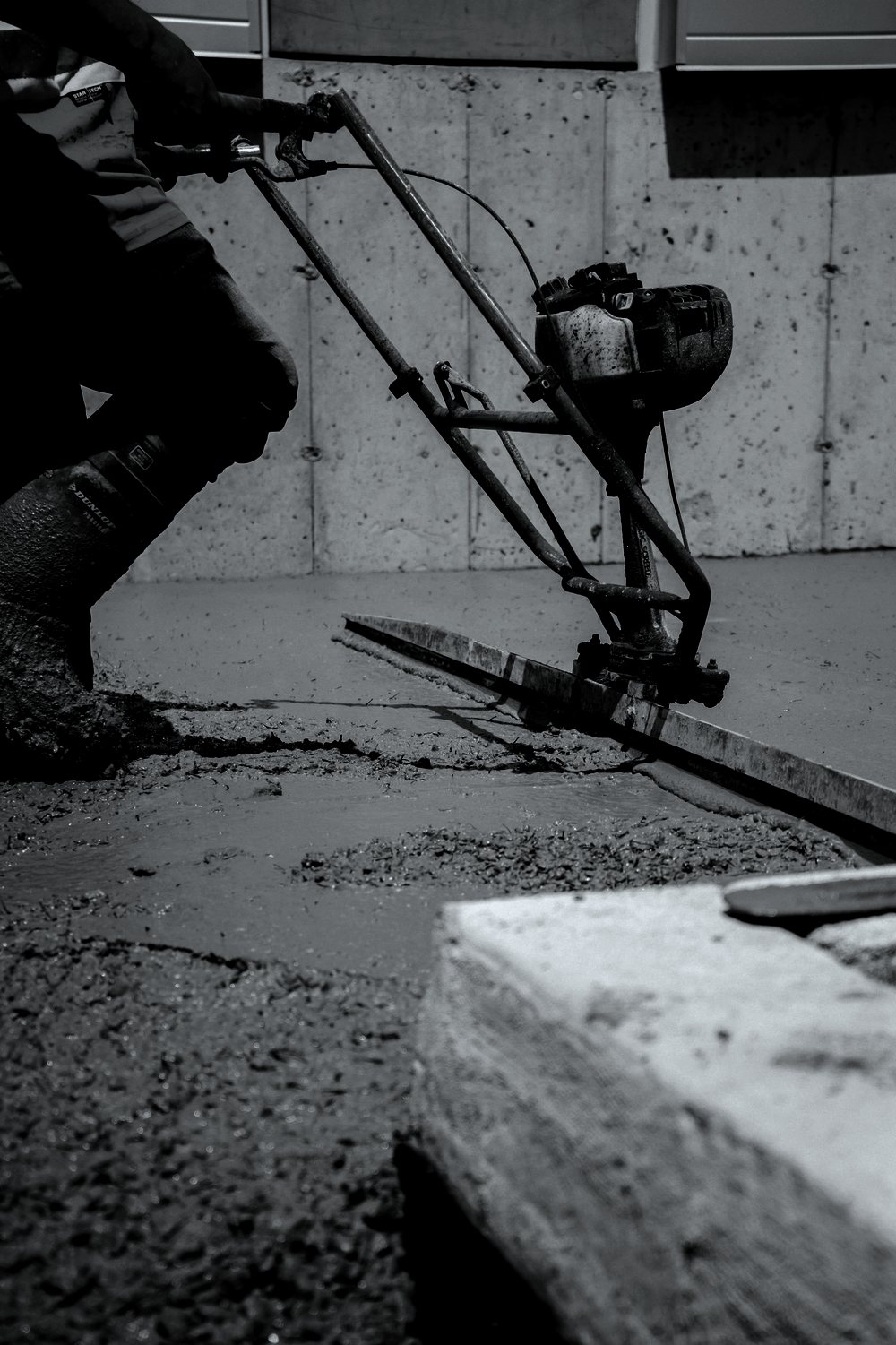 A worker running a walk behind power screed across a fresh slab beside a formed concrete wall.